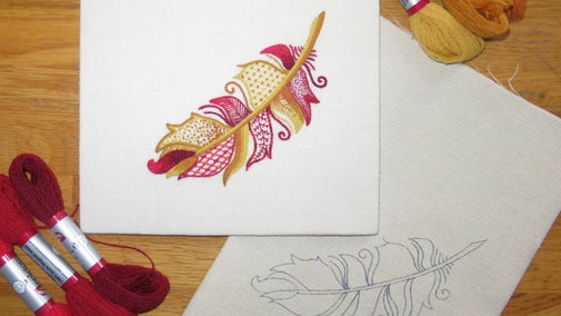 The image depicts an intricate embroidery design of a feather, predominantly featuring shades of red, yellow, and orange thread. The design includes various textures and patterns, such as dots, swirls, and grids, adding depth and complexity to the feather. To the left of the finished embroidery, there are red threads neatly arranged, and to the right, there are more orange and yellow threads. Additionally, a fabric with an outline of the same feather design, yet to be embroidered, is visible in the lower right corner, showing the preparatory sketch. The setup suggests a work-in-progress embroidery project.